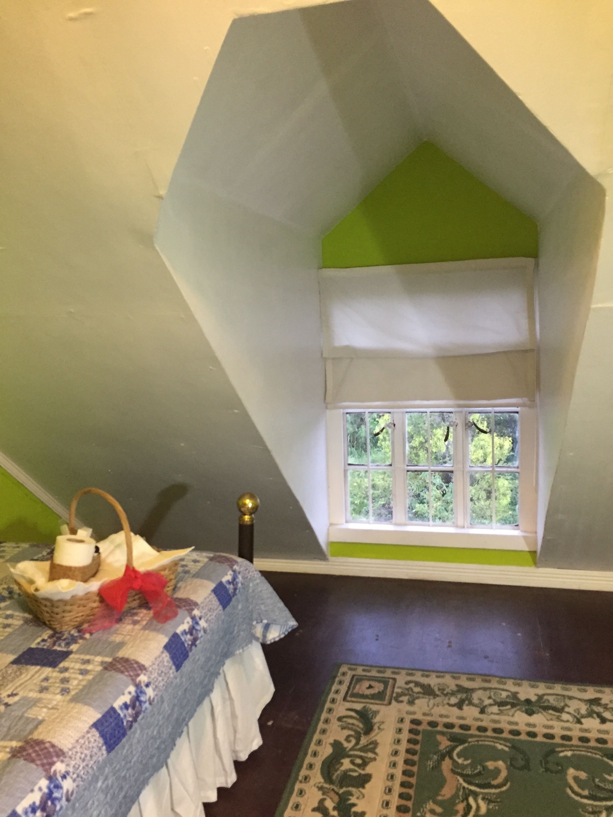 A cozy attic room with a sloped ceiling, a small window with a view of greenery, a quilted bedspread, and a patterned rug.