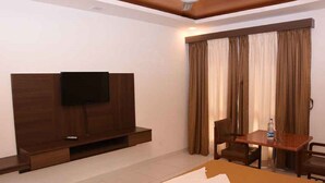 A hotel room with a flat-screen TV mounted on a wooden entertainment unit, a small table with a chair, and a window with curtains.