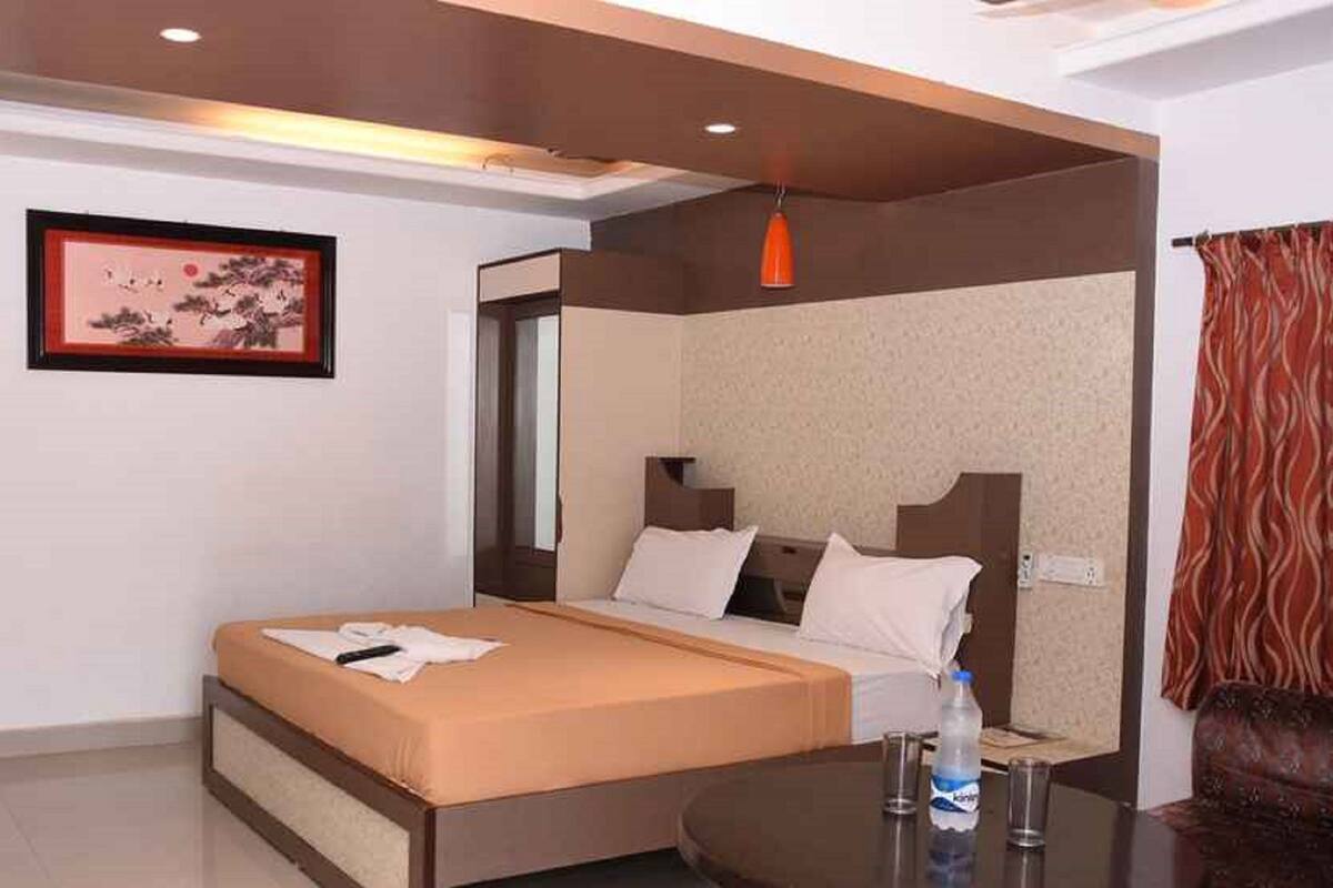 A hotel room with a bed, a small table with glasses and a water bottle, a framed picture on the wall, and a red curtain.