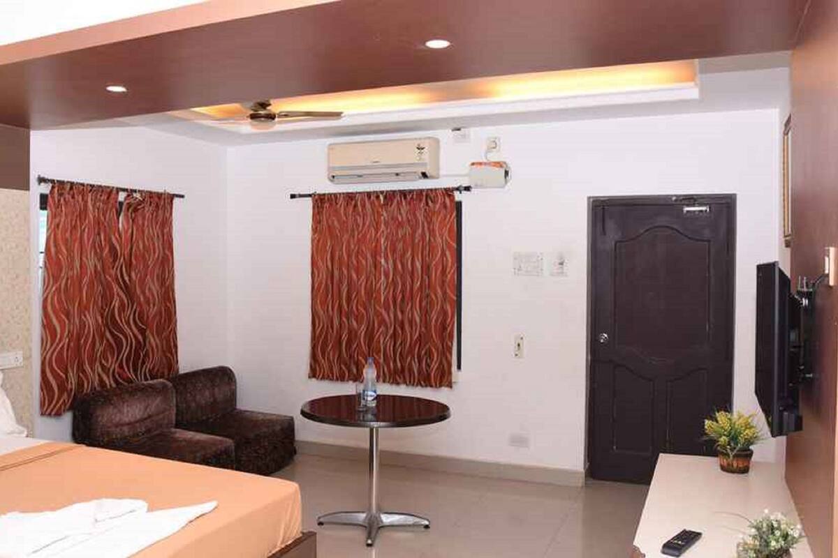 A hotel room with a bed, two armchairs, a small round table, an air conditioning unit, a door, and a television.