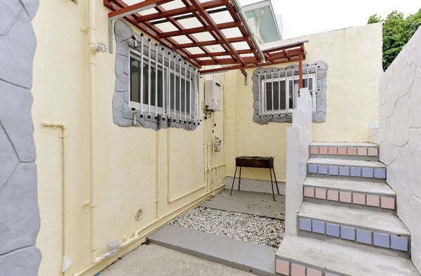 Un espacio exterior pequeño y cerrado con piso de concreto, una parrilla metálica y una pérgola de madera.