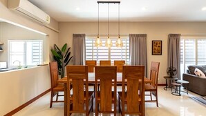 A dining area with a wooden table and chairs, a hanging light fixture, a window with curtains, and a plant.