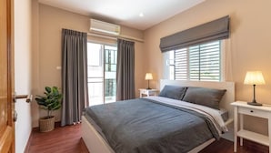 A bedroom with a large bed, a window with blinds, a bedside table with a lamp, and a potted plant.