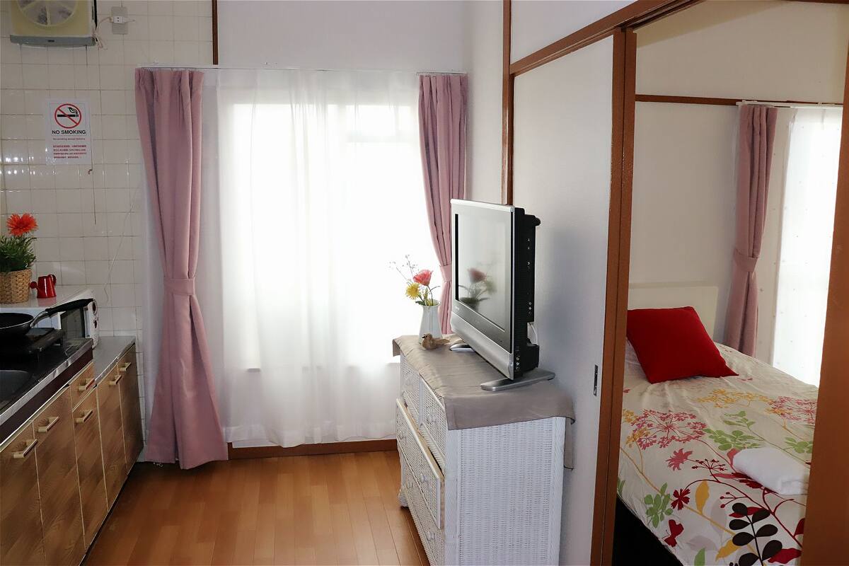 A room with a bed, a TV on a stand, a kitchen area with a stove and a sink, and a window with curtains.