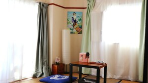 Une pièce avec des tatamis, une petite table sur laquelle se trouvent une boîte rose et une télécommande, un coussin bleu et un tableau accroché au mur.