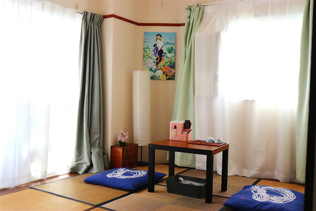 A room with tatami mats, a small table with a pink box and remote, a blue cushion, and a painting on the wall.
