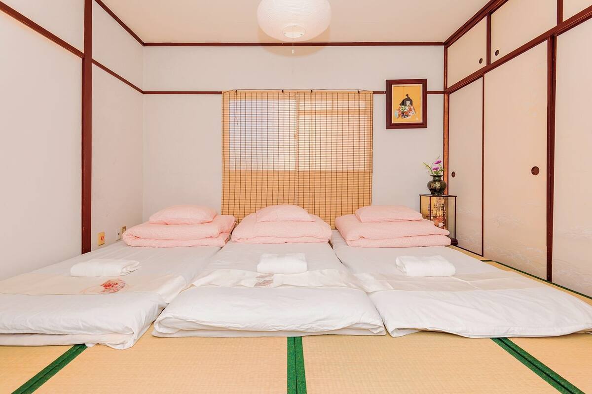 A traditional Japanese-style room with tatami flooring, a futon bed with pink and white bedding, a shoji screen, and a small shelf with a vase.