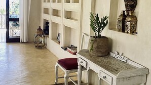 A room with a white wooden bench, a red cushioned chair, a decorative plant, and a wall-mounted shelf with books and a lantern. - Monkey Beach House, Beachfront, Ocean Views,  Shared Pool, Staff Provided (Diani Beach)