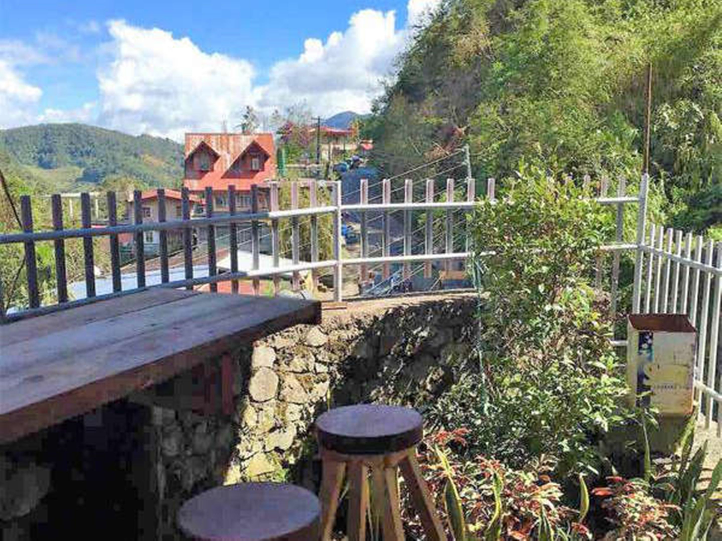 A rustic stone bridge with a wooden deck and metal railing, overlooking a lush valley with houses and greenery.