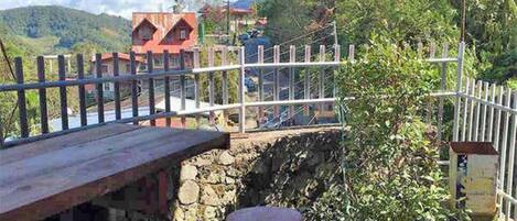 A rustic stone bridge with a wooden deck and metal railing, overlooking a lush valley with houses and greenery.