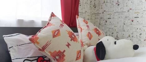 Una cama con un cojín de Hello Kitty, un peluche de Snoopy y una pared con estampado.