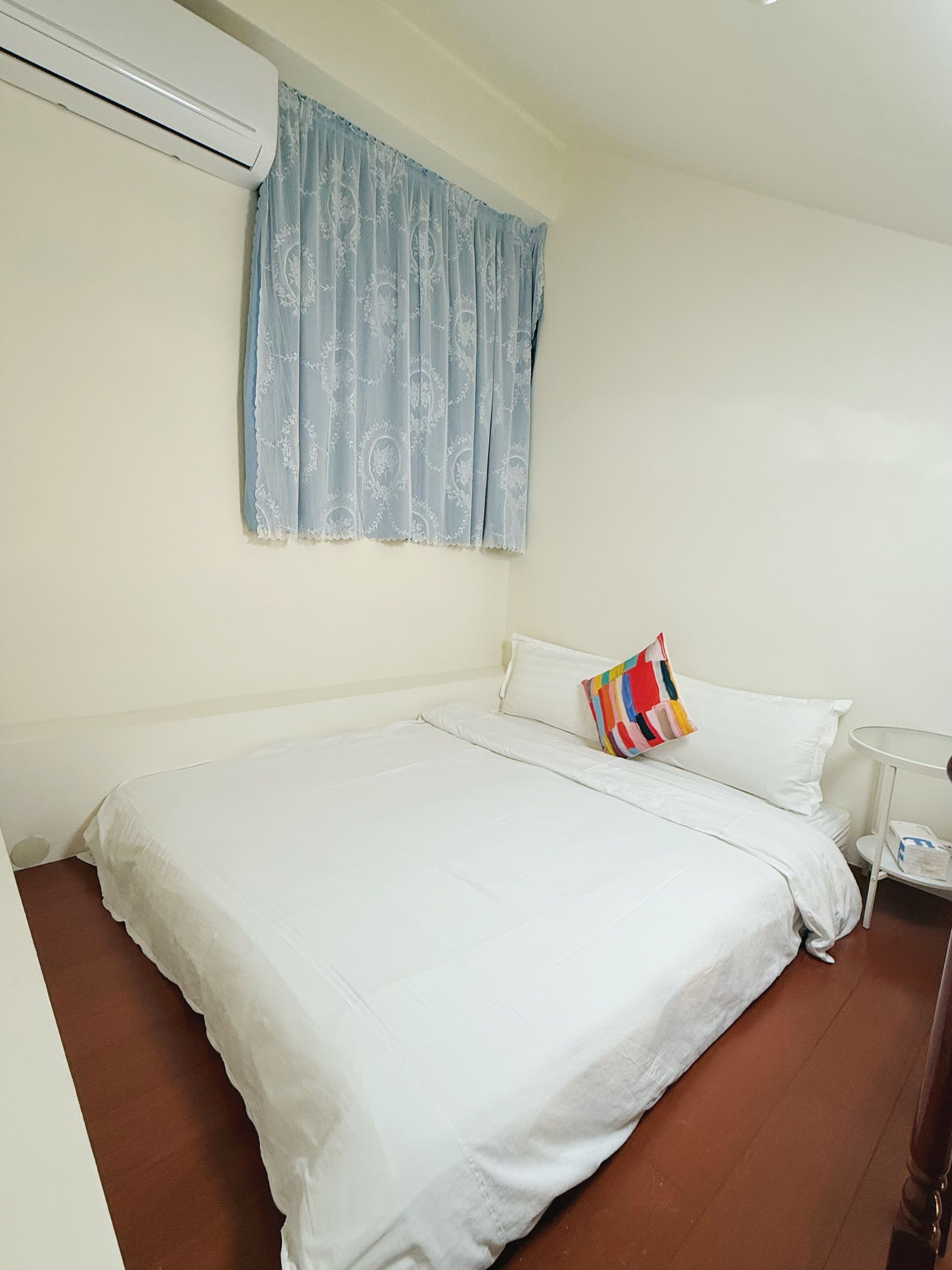 Habitación de hotel con una cama individual, una mesita de noche pequeña, un almohadón blanco y uno colorido, y una ventana con cortina azul.
