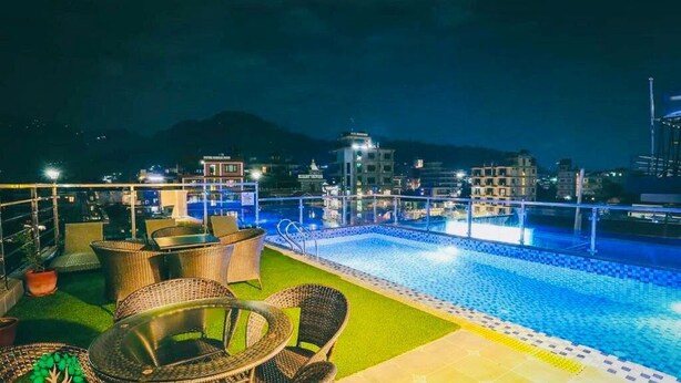Un área de piscina en la azotea con mobiliario de mimbre y vista a la ciudad de noche.