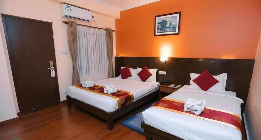 A hotel room with two beds, a wooden floor, a door, an air conditioning unit, and a painting on the wall.