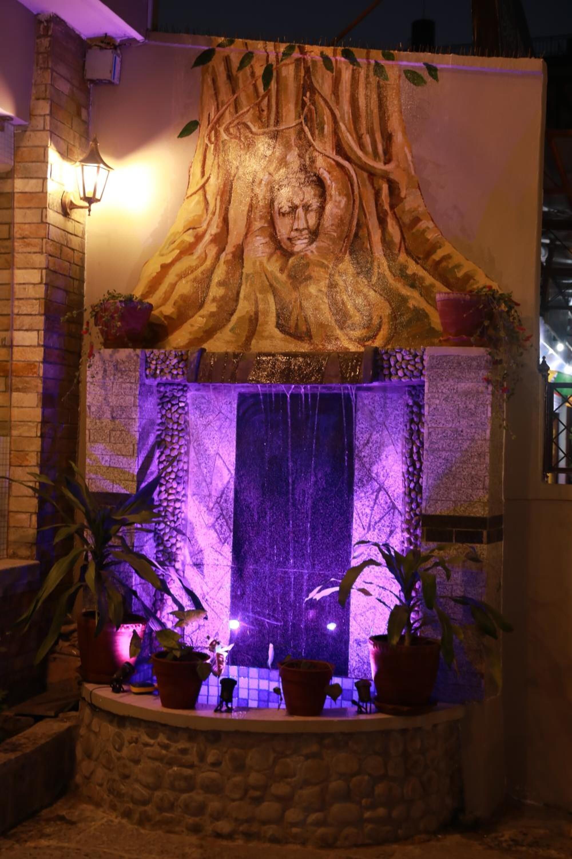 A stone fountain with a carved tree trunk and a Buddha statue, surrounded by potted plants.
