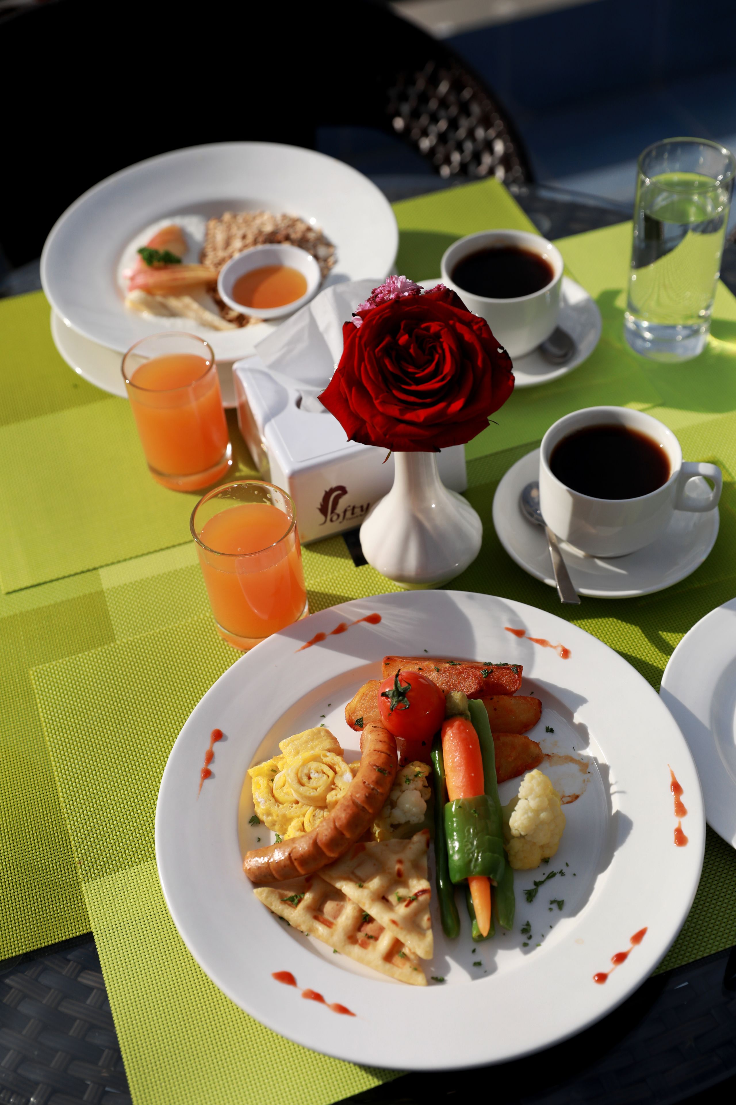 Une table dressée avec des assiettes remplies de nourriture, des verres de jus de fruits et un vase contenant une rose rouge.