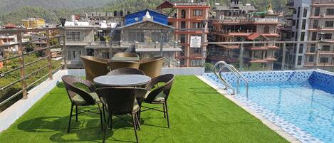 A rooftop pool with artificial grass and wicker furniture, surrounded by hotel buildings with visible signage.