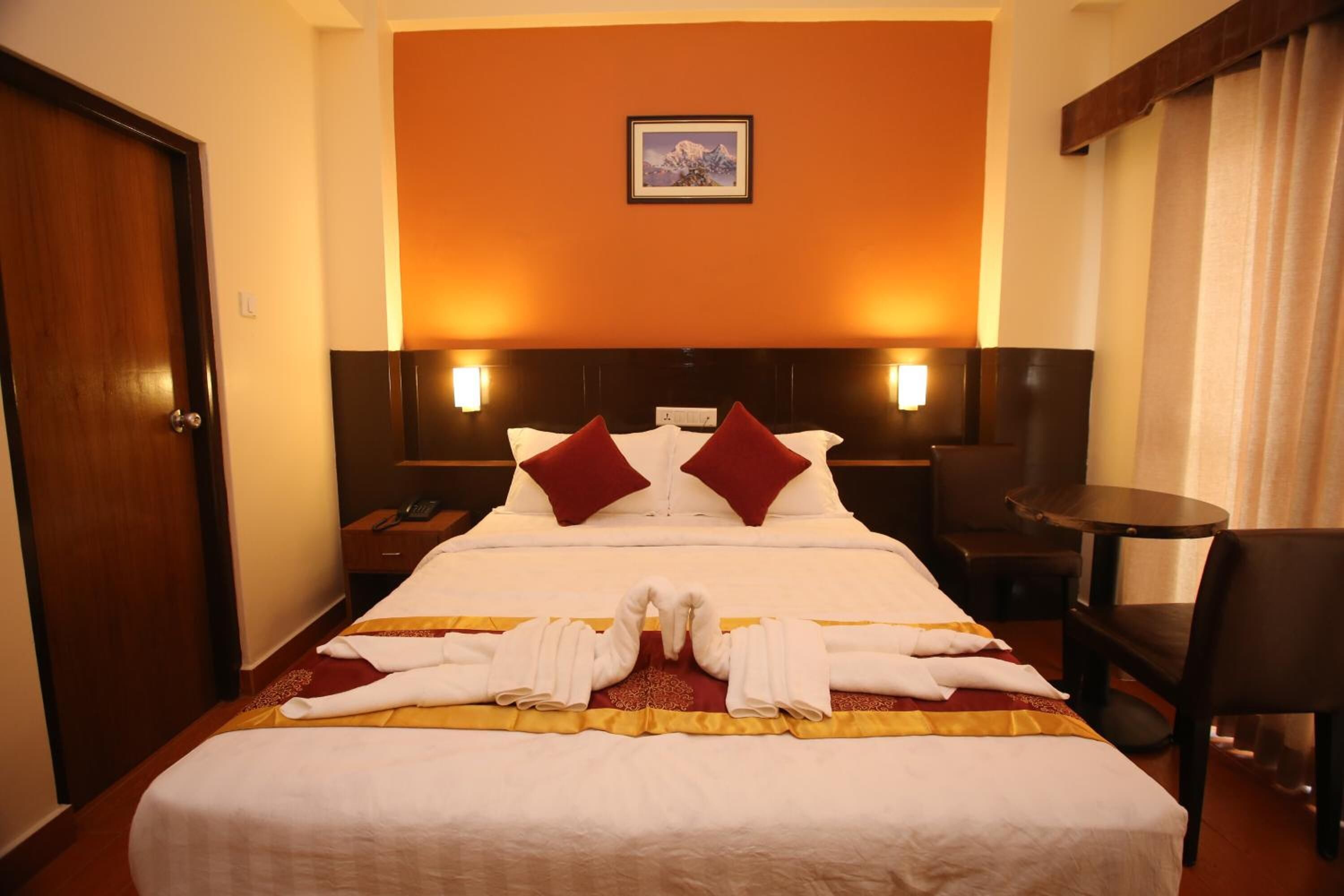 A hotel room with a large bed, two red pillows, a headboard with wall-mounted lights, a wooden door, a small table with a chair, and a framed picture on the wall.