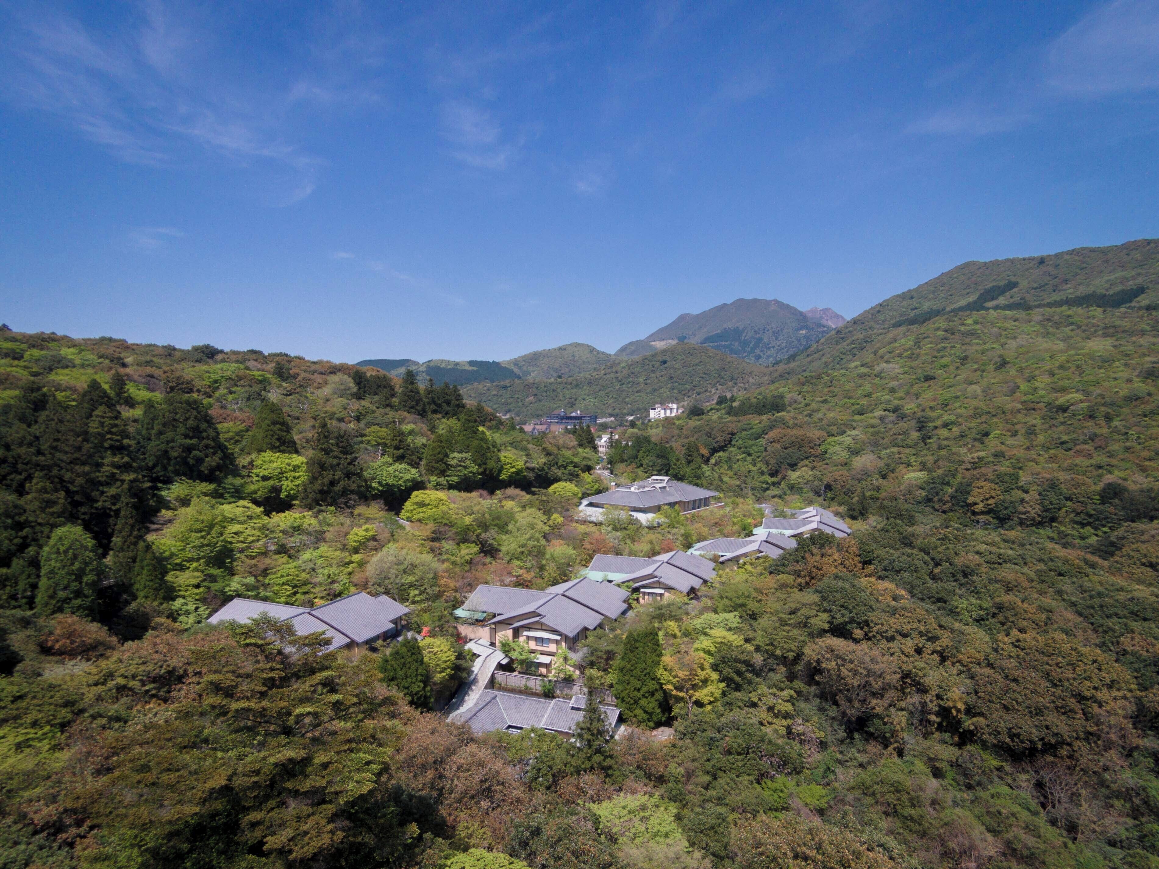 A cluster of traditional-style buildings with sloped roofs, surrounded by dense forest and hills in the background.