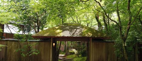 A traditional wooden gazebo with a tiled roof, surrounded by lush greenery and a stone pathway leading to it.