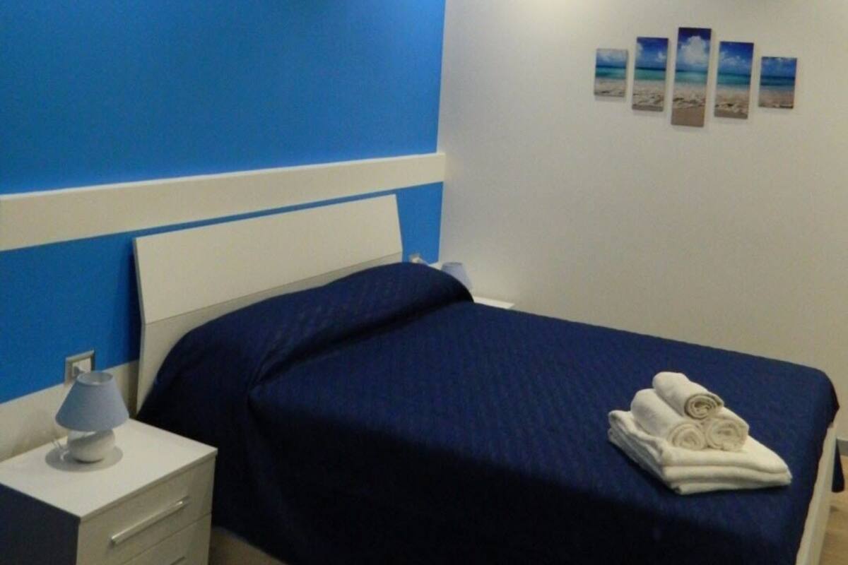 Ein Schlafzimmer mit einem Bett, einem Nachttisch, einer Lampe und einer Wand mit vier gerahmten Bildern.