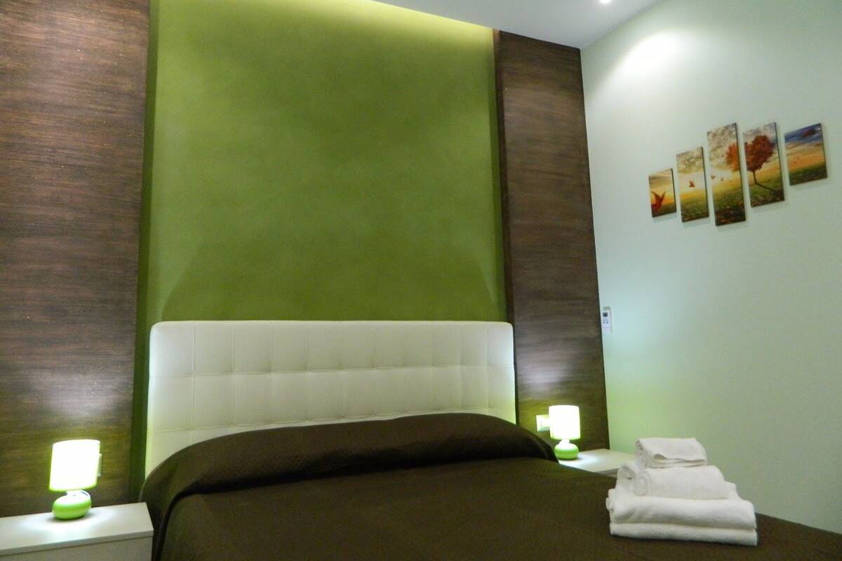 Ein Hotelzimmer mit einem großen Bett, Wandverkleidungen in Grün und Braun, einem weißen Kopfteil sowie einem Nachttisch mit Lampe und Handtüchern.