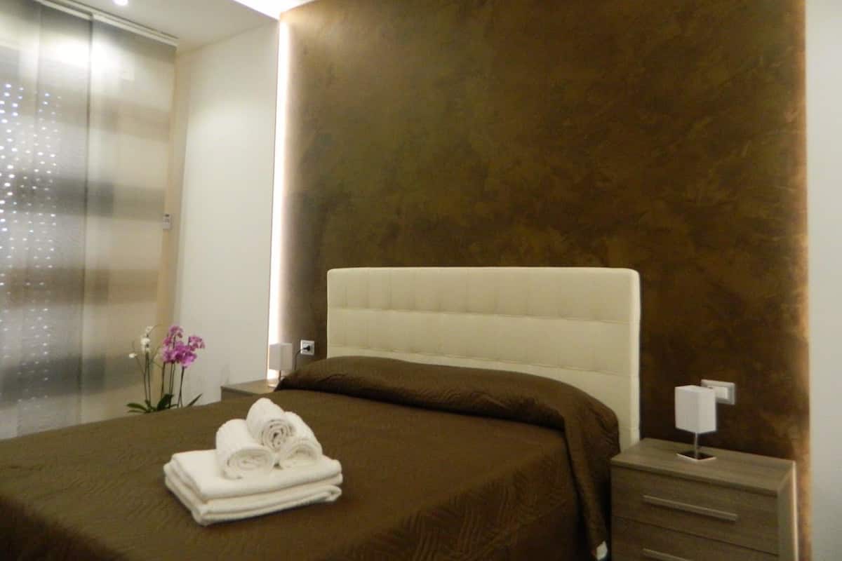 Ein modernes Hotelzimmer mit einem großen Bett, einem Nachttisch, einer Lampe und einer Vase mit Blumen.