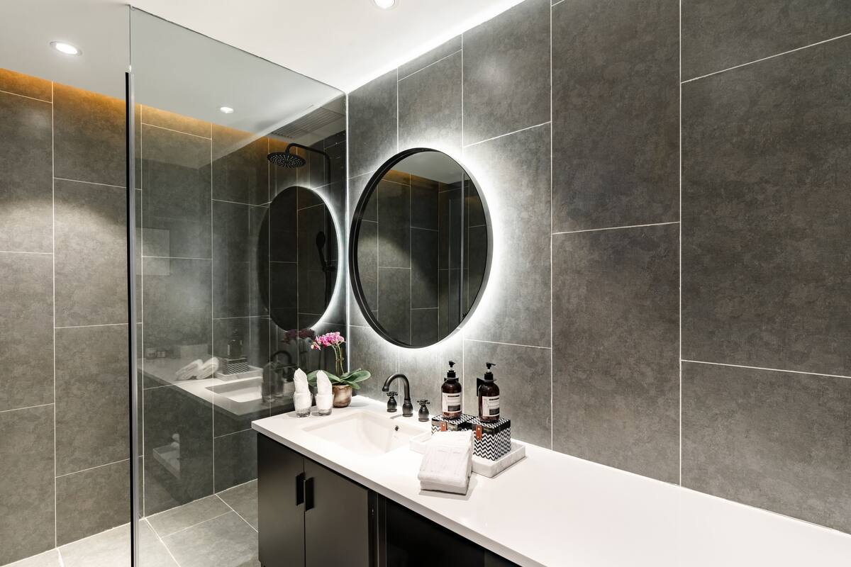 A modern bathroom with a glass shower enclosure, a white sink with a black soap dispenser, and a large mirror above the sink.
