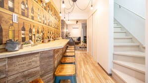 A bar area with wooden bar stools, a counter displaying wine bottles, and a staircase to the right. - Bar Hostel Shikanzima (Osaka)