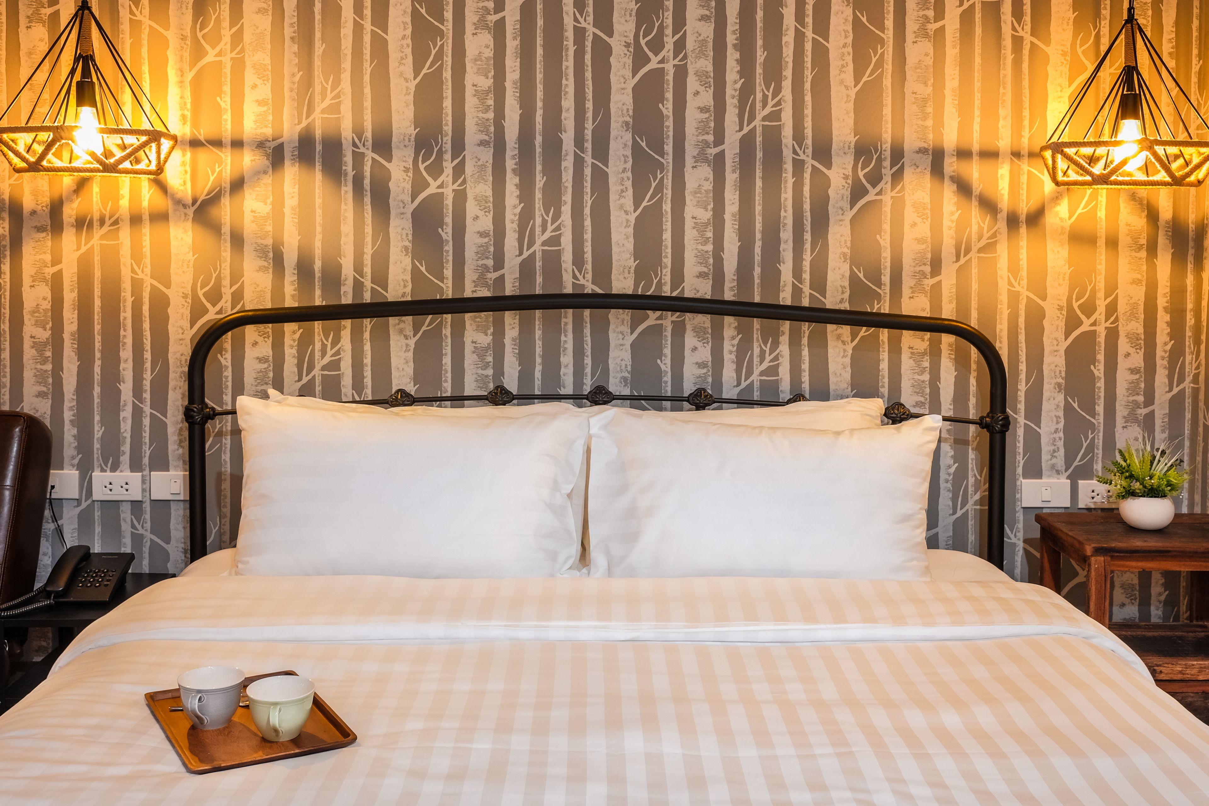 Ein ordentlich bezogenes Bett mit weißen Leinentüchern, ein Tablett mit zwei Tassen und ein kleiner Topfpflanzen auf einem Holznachttisch.