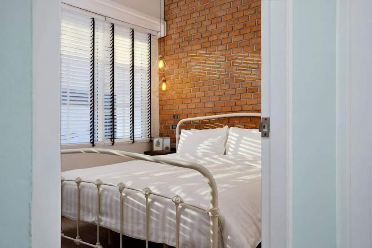 Ein Bett mit Metallgestell, eine Ziegelwand und ein Fenster mit Jalousien.