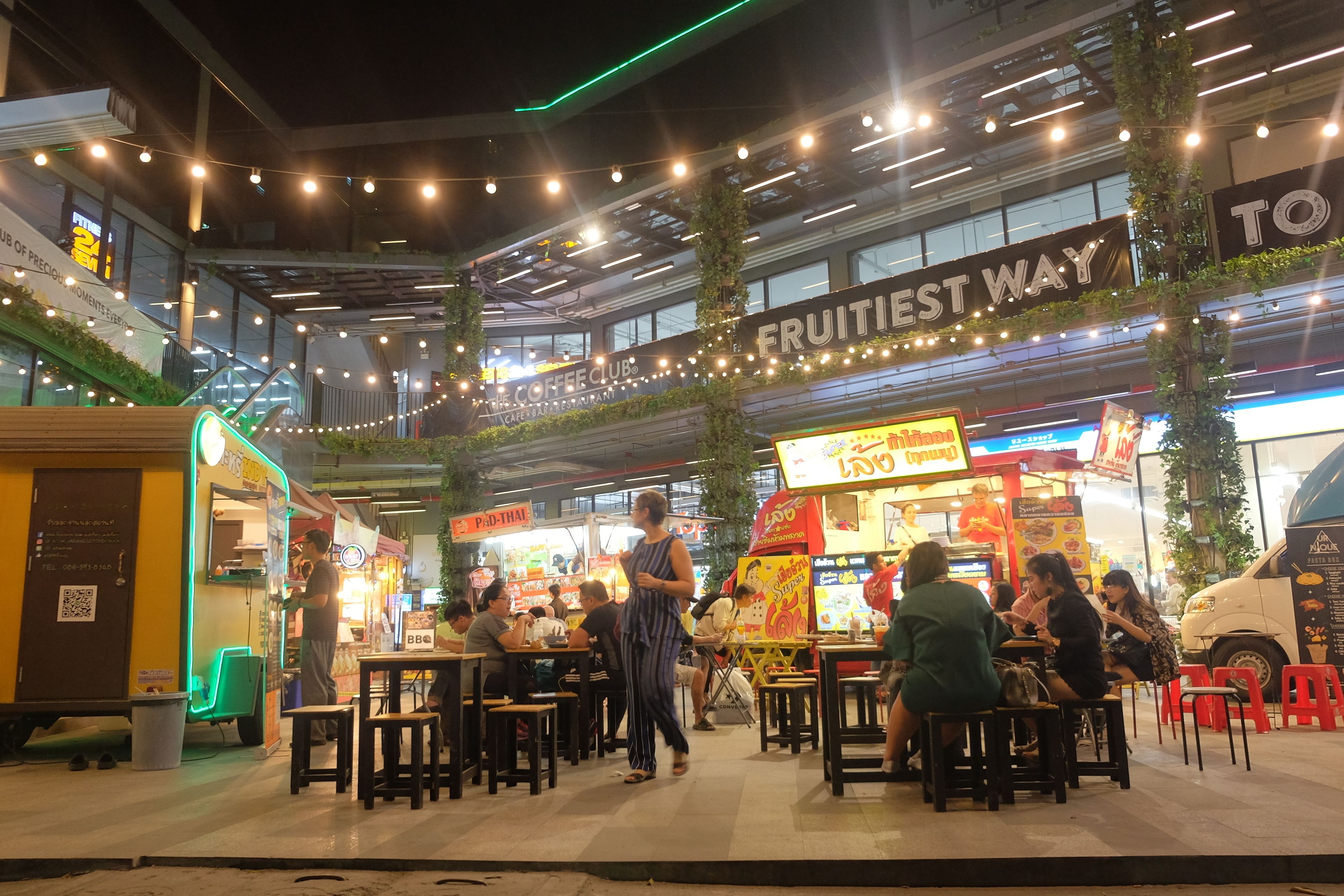An outdoor dining area with tables and chairs, a sign reading "FRUITIEST WAY", and a food truck in the background.