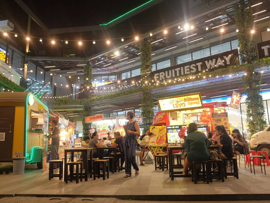 An outdoor dining area with tables and chairs, a sign reading "FRUITIEST WAY", and a food truck in the background.