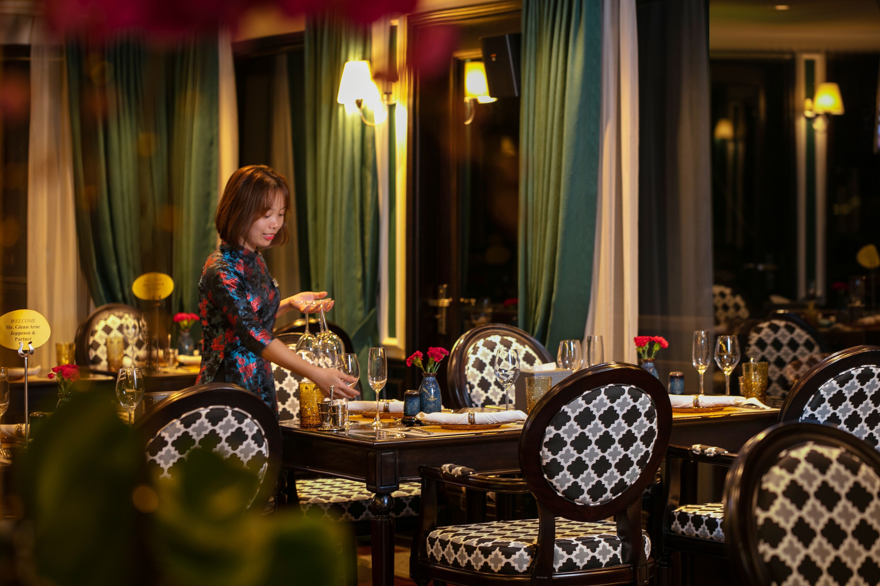 Une salle à manger où une personne est en train de mettre la table. La pièce est décorée de chaises et de tables à motifs, et on y trouve des rideaux et des lampes.