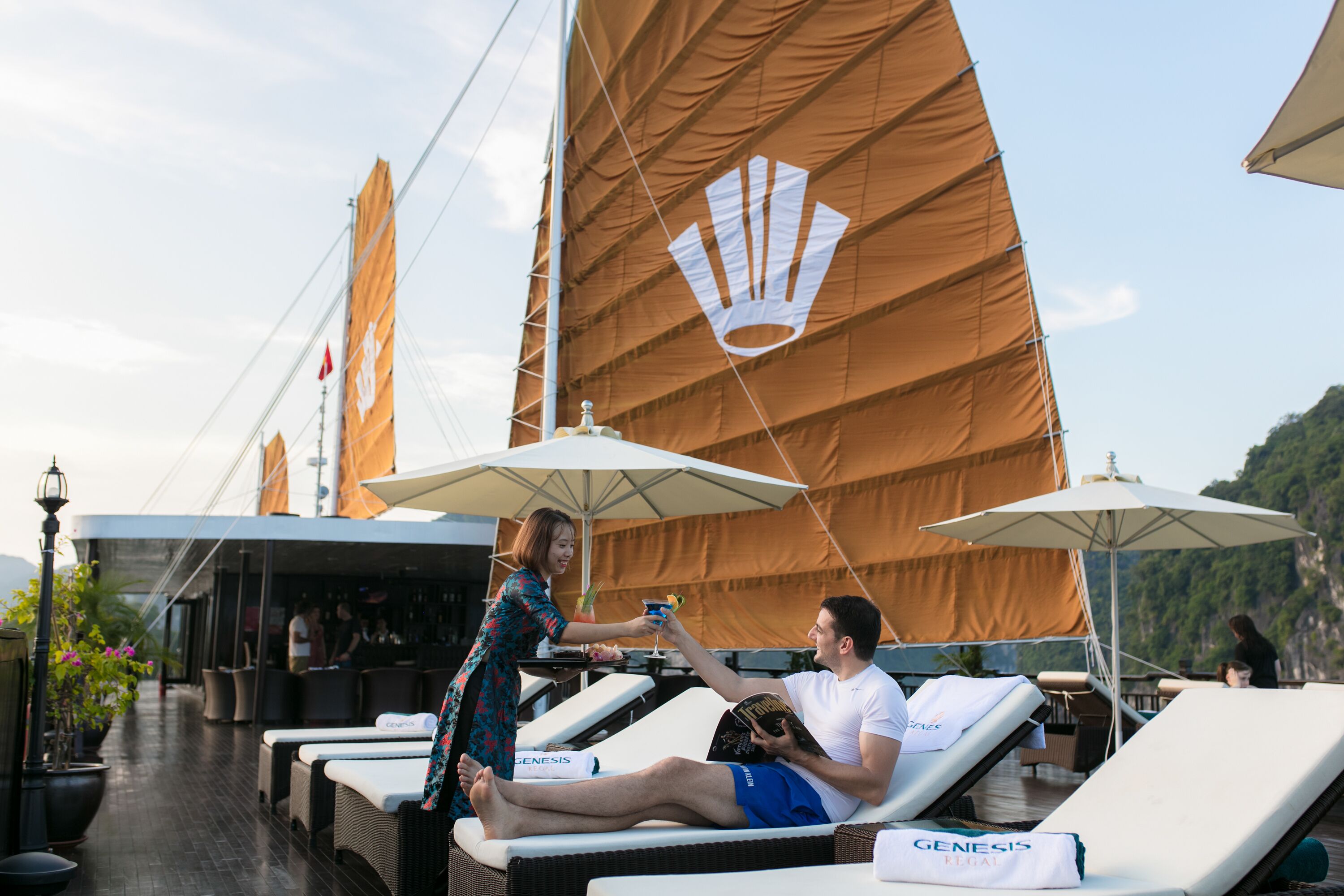 A couple relaxing on sun loungers with umbrellas, a bar area, and a view of a mountainous landscape.