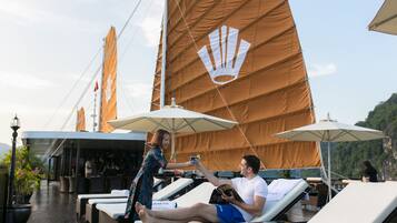 A couple relaxing on sun loungers with umbrellas, a bar area, and a view of a mountainous landscape.