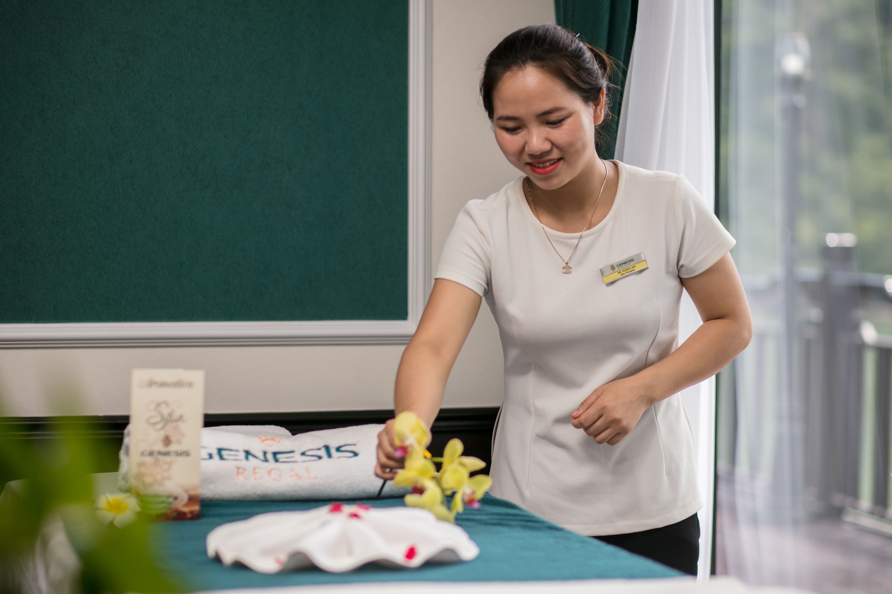 A person in a white shirt arranging flowers on a bed with a green bedspread and a white pillow labeled "GENESIS REGAL".