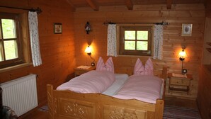 A wooden cabin bedroom with a double bed, two bedside tables, a nightstand, a window with curtains, and a radiator.