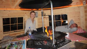 A wooden cabin interior with a central fireplace, a table with a checkered tablecloth, and a person lying on a couch.