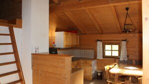 A wooden cabin interior with a kitchen area, wooden dining table, chairs, and a window with curtains.
