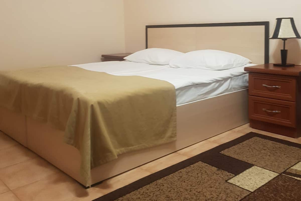 A single bed with a beige bedspread, a wooden nightstand with a lamp, and a patterned rug on a wooden floor.
