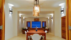 A dining area with a wooden table and chairs, a hanging light fixture, and a view of the sea through large windows. - Four Bedroom Ecoluxe Haven in Treasure Beach with Pool (Treasure Beach)