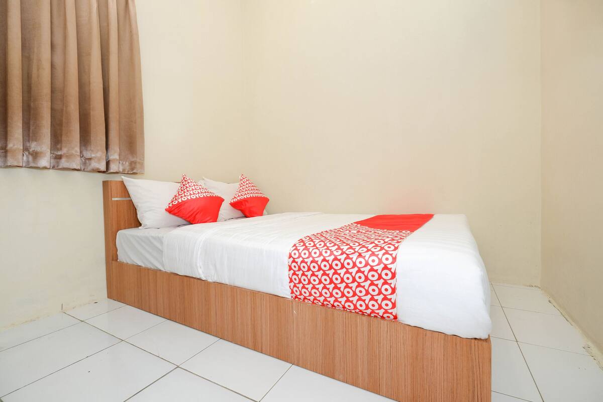 Uma cama de solteiro com estrutura de madeira, roupa de cama branca e um cobertor estampado em vermelho e branco.