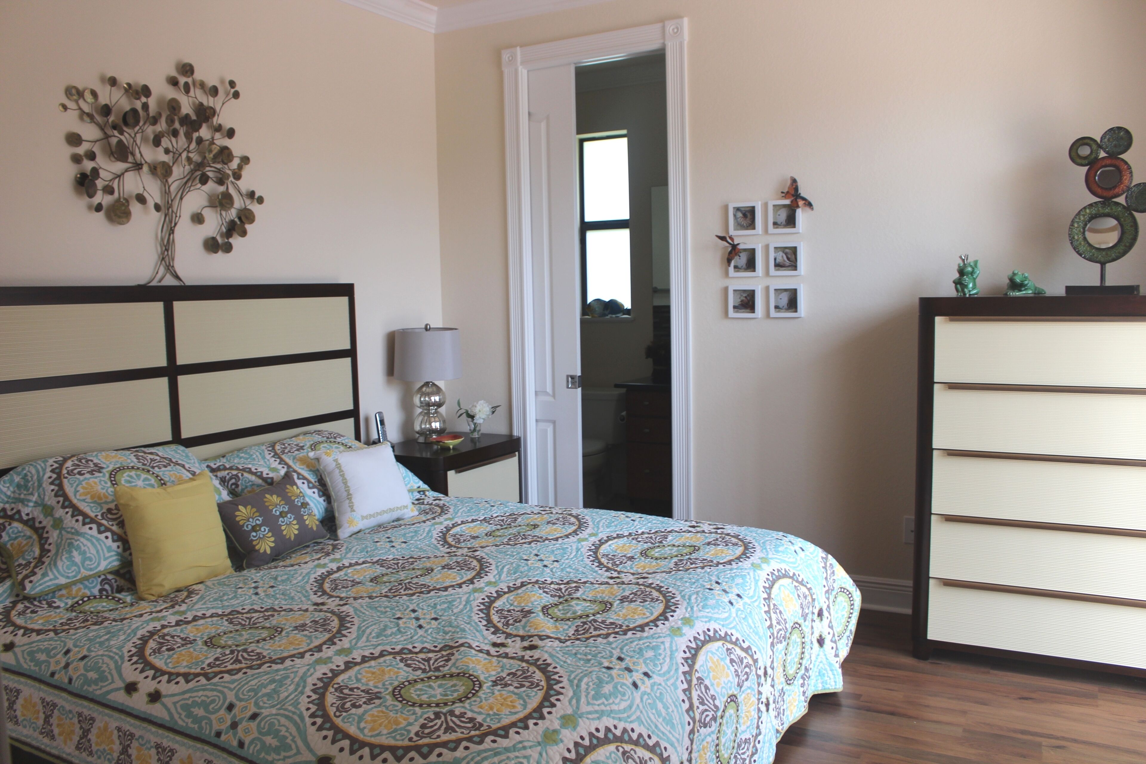 Une chambre à coucher avec une couvre-lit à motifs, une table de chevet avec une lampe, une commode et un arbre décoratif peint sur le mur.
