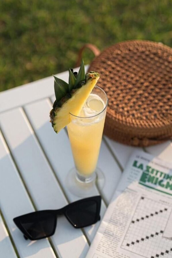 A glass of a yellow beverage with a pineapple garnish, sunglasses, and a menu on a white table.