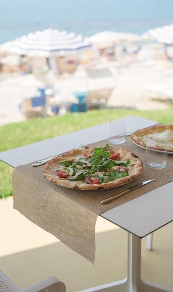 Una mesa con pizza y un vaso de agua, con vista a una playa con sombrillas.