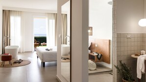 Una habitación de hotel moderna con una cama grande, una zona de estar con una silla y reposapiés, vistas al exterior y un baño con ducha y lavamanos.
