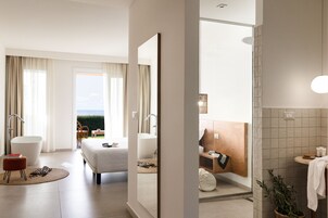 Una habitación de hotel moderna con una cama grande, una zona de estar con una silla y reposapiés, vistas al exterior y un baño con ducha y lavamanos.