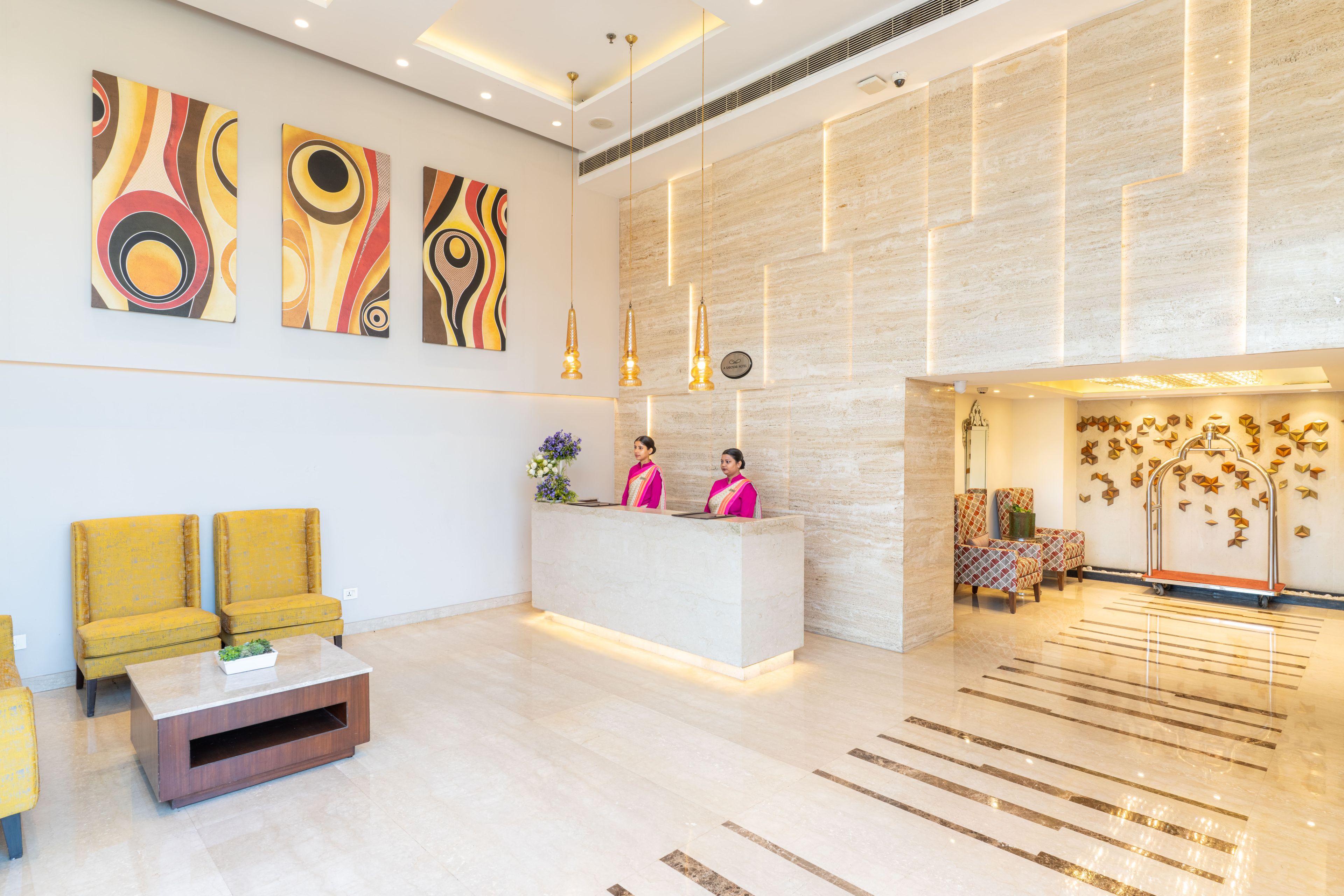 Een hotellobby met een receptiebalie, twee receptionisten, een gele bank, een bijzettafeltje en abstracte wanddecoratie.