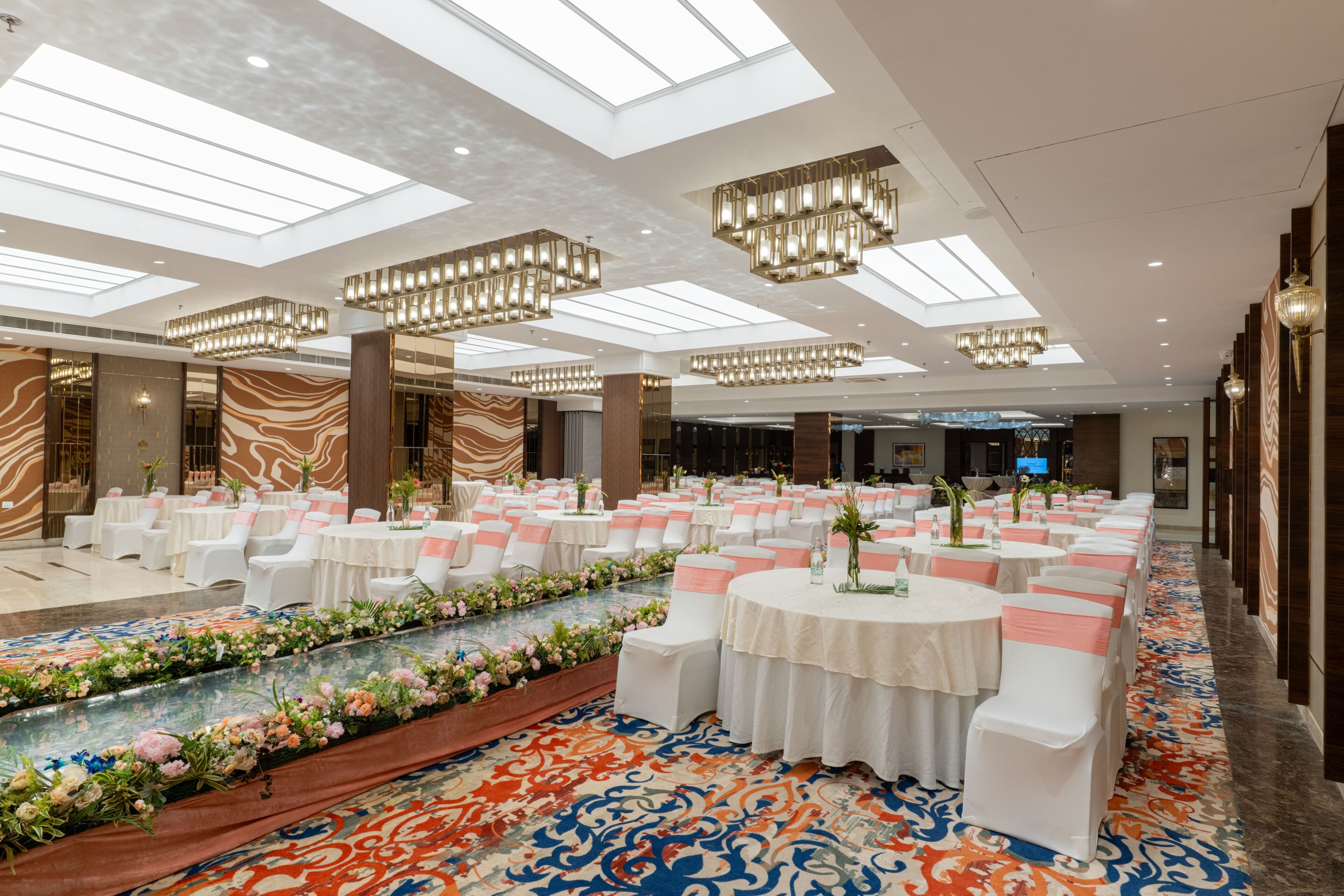 A spacious banquet hall with round tables, white chairs, and a decorative floral arrangement along the walkway.